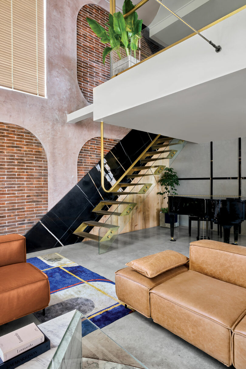 Interior of a modern living room featuring a black staircase with gold accents, a grand piano, brown leather seating, and a colorful area rug against a backdrop of brick and plaster walls.