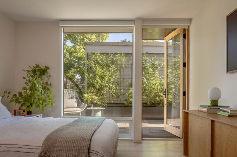 Bedroom with a double bed, large window with a view of a garden patio, and an open glass door leading outside. A plant, side table with books, and a small lamp are visible.