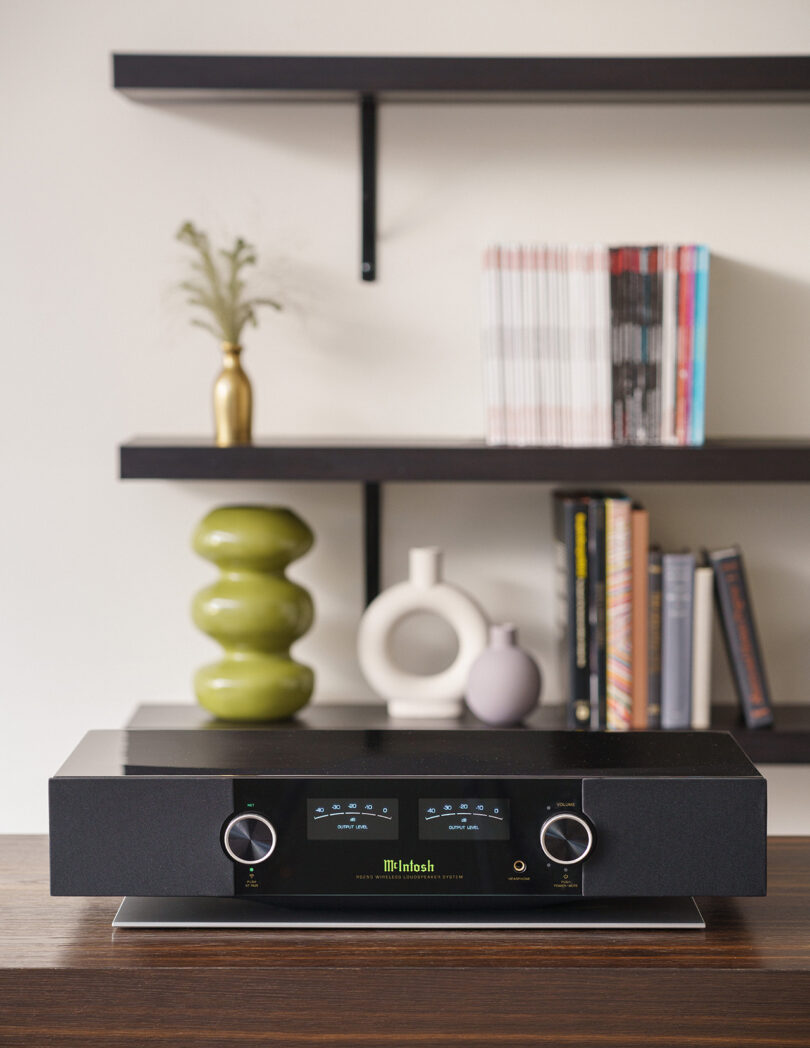 A stereo amplifier with dual dials is placed on a wooden surface. In the background are shelves with decorative items and books.
