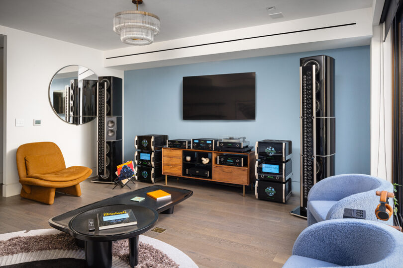 Modern living room with a wall-mounted TV, large speakers, and wooden media unit. It features a brown chair, round mirror, and two blue armchairs on a patterned rug. Ceiling light fixture above.