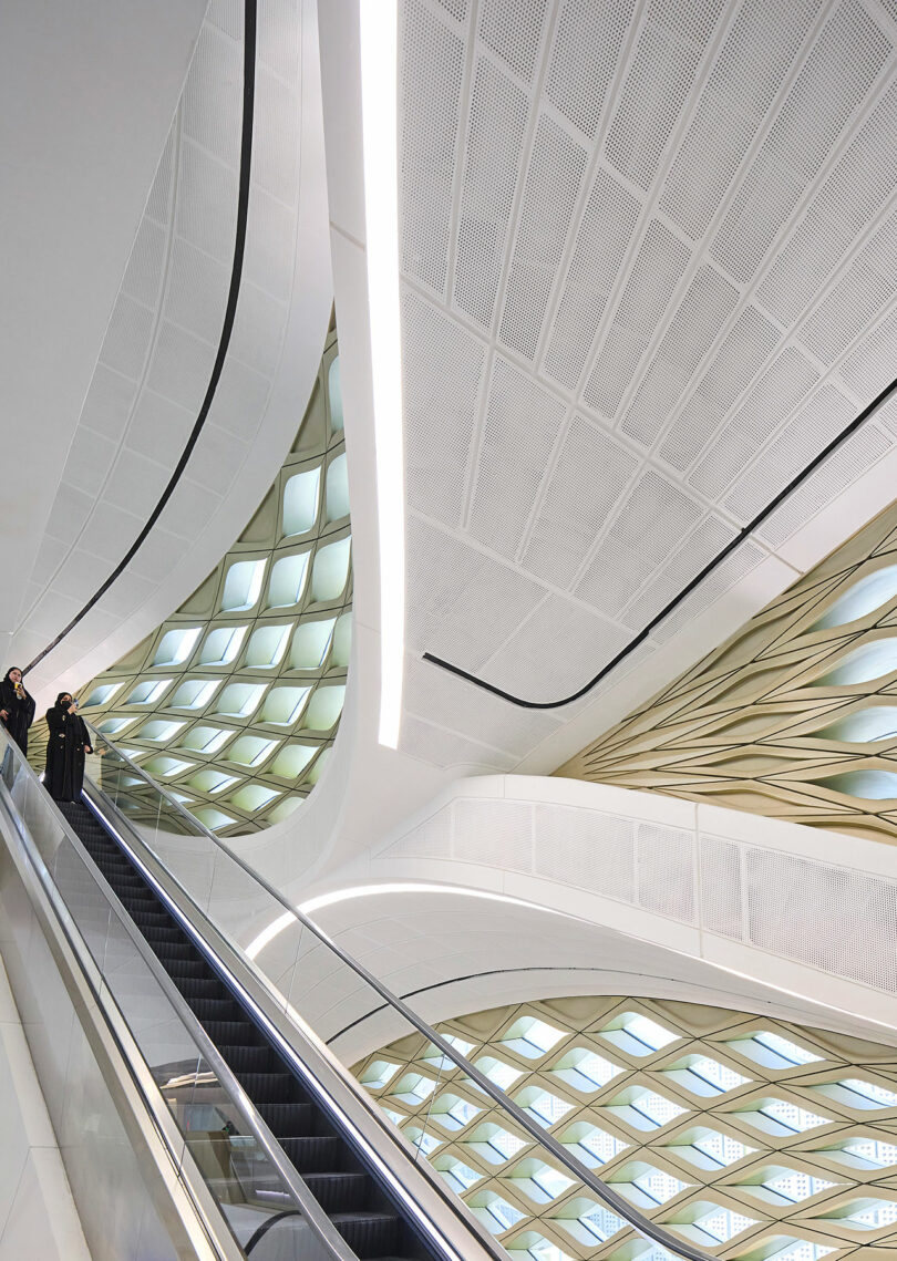 People ascend an escalator inside a modern building with a futuristic design featuring geometric skylights and curved surfaces.