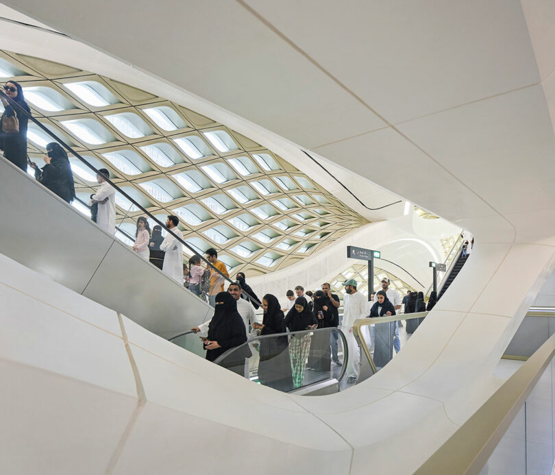 People riding escalators in a modern, geometric-styled building with a lattice-patterned ceiling and white structural elements.
