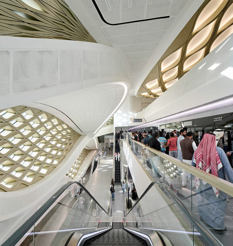 Futuristic train station interior with modern architectural design. People are using escalators and lining up at a platform.