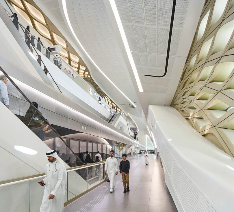 Modern interior of a transport hub with sleek, white architecture. People walk along a corridor and use escalators to various levels, surrounded by geometric patterns and bright lighting.