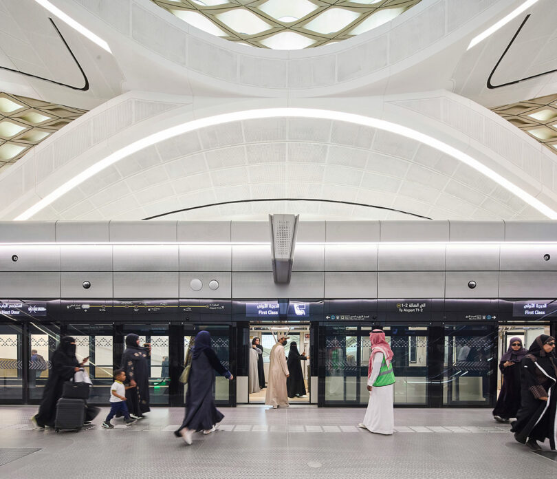 People walking and standing at a modern, spacious subway station with geometric ceiling design and sliding glass doors.
