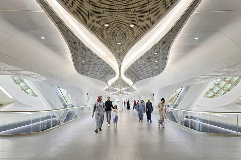 Spacious futuristic walkway with a curvy ceiling design. Several people are walking along the path, surrounded by sleek architecture.