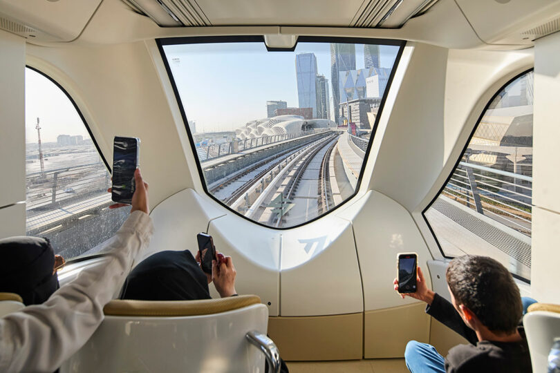 Passengers taking photos from inside a moving train, with a large front window view showing tracks and distant city skyscrapers.