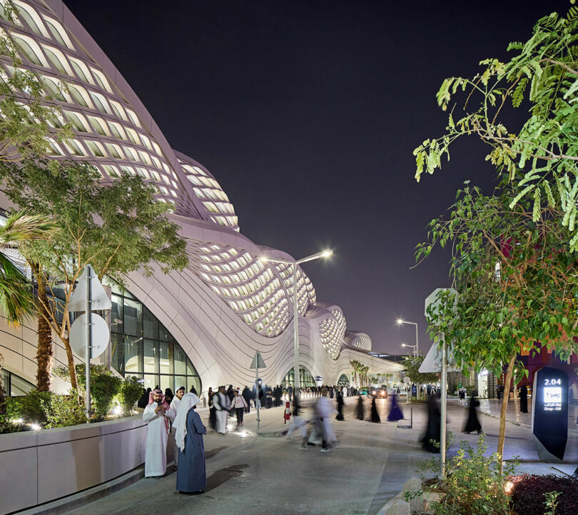 Night scene of an outdoor modern architectural space with curved structures, trees, and people walking around.