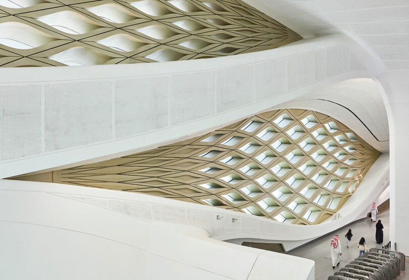 Interior of a modern architectural space with geometric patterns on walls and ceiling. People in traditional attire walk through the area.