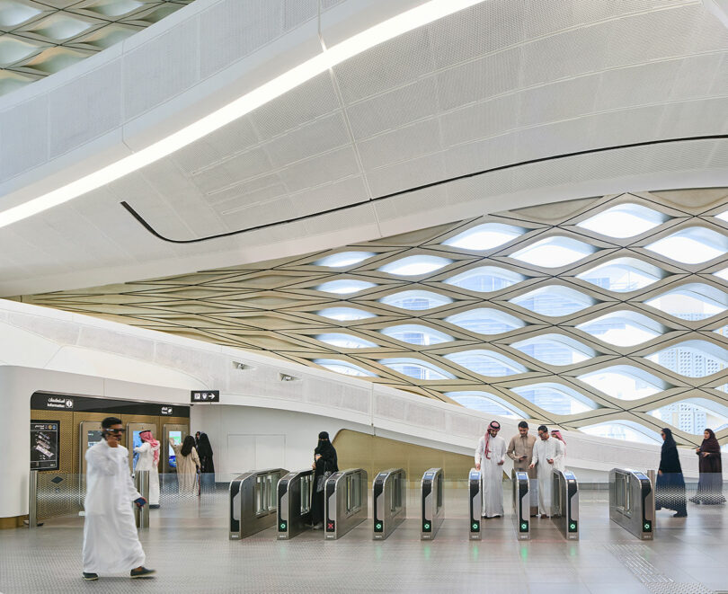 A modern metro station entrance with a geometric ceiling design. People are passing through ticket barriers. The station has a spacious and bright interior.