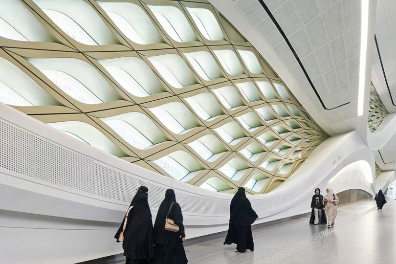 People walk through a modern architectural space featuring a curving, lattice-patterned ceiling with large windows.
