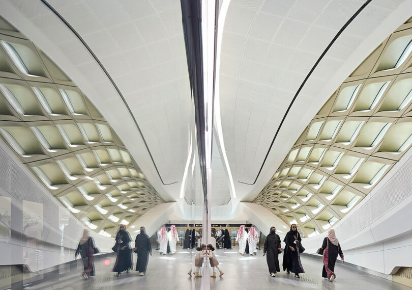 Futuristic train station interior with large, curved ceiling and patterned walls. People walk along the platform, some in traditional attire, reflecting on the polished floor.