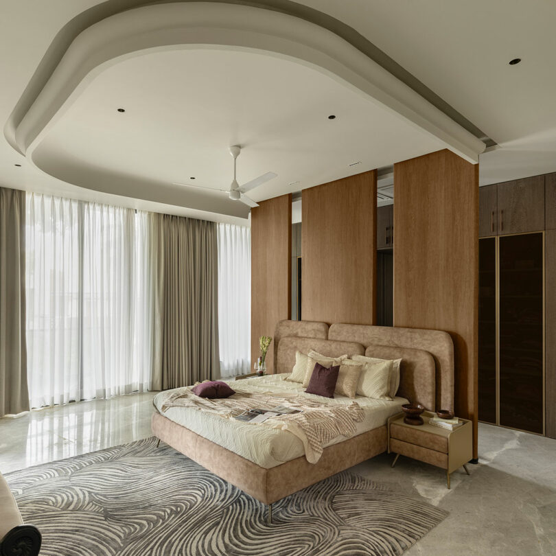 Modern bedroom with a large bed, neutral-toned bedding, wooden partition, abstract rug, ceiling fan, and floor-to-ceiling curtains allowing natural light.
