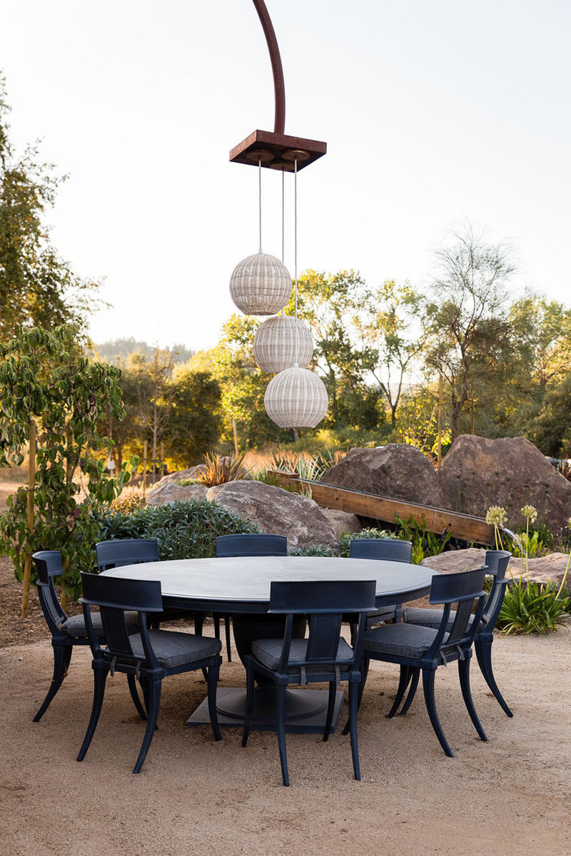 This elegant outdoor dining area features a round table surrounded by comfortable, contemporary chairs. The space is framed by rugged rock landscaping with soft lighting to enhance the tranquil atmosphere. The combination of natural elements and contemporary design creates a sophisticated yet inviting environment, making it an ideal spot for alfresco meals and gatherings.