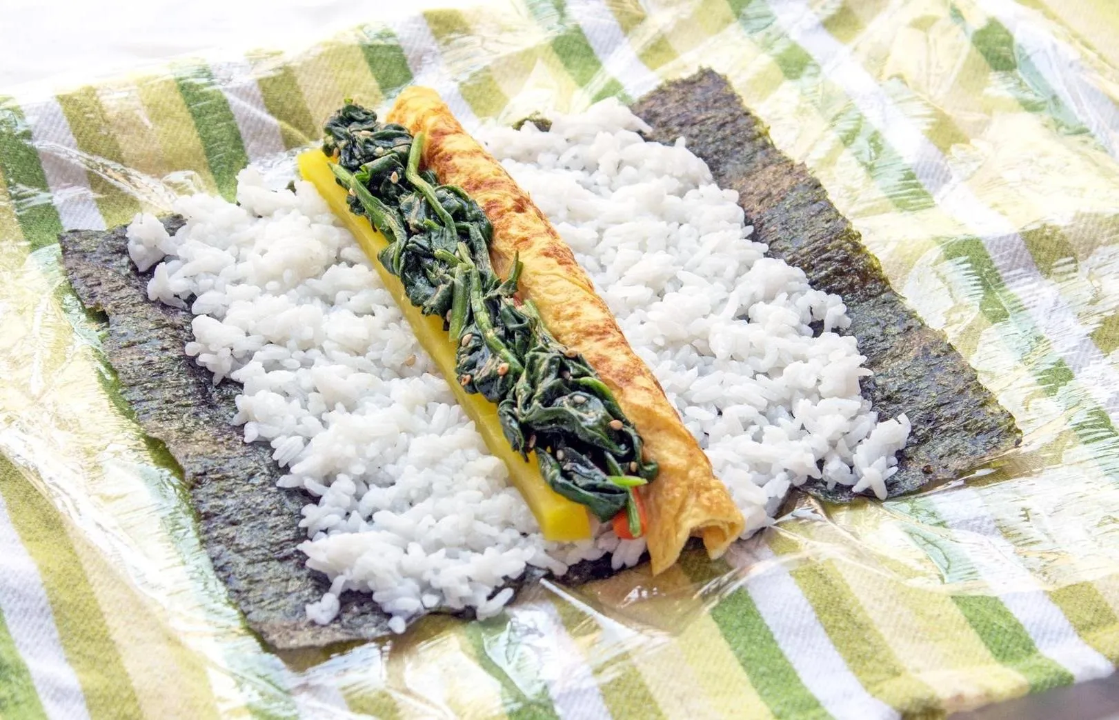 Vegetarian sushi roll ingredients – rice, tamago, spinach, burdock root, and pickled daikon radish – on a sheet of seaweed on a plastic-wrap-covered tea towel.
