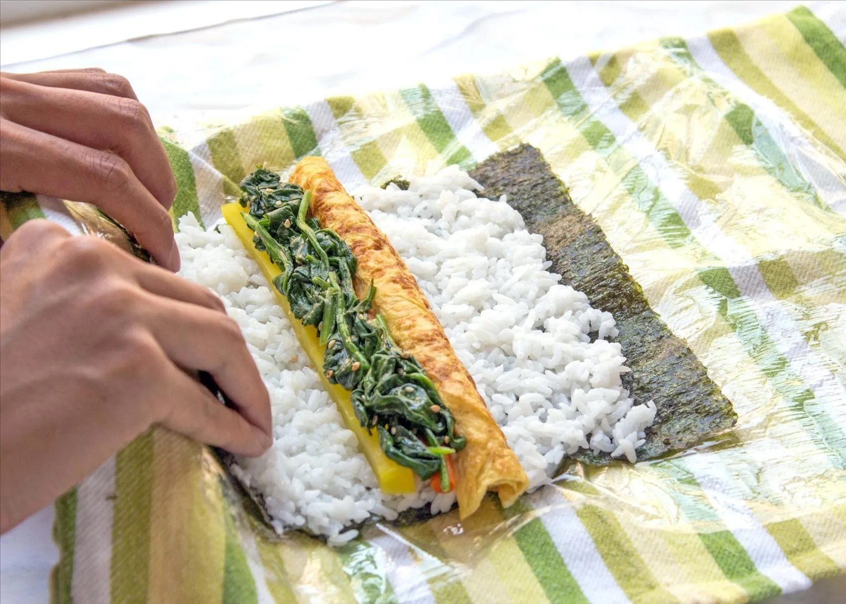 Vegetarian sushi rolls being rolling with sushi ingredients – rice, tamago, spinach, burdock root, and pickled daikon radish – on a sheet of seaweed on a plastic-wrap-covered tea towel.