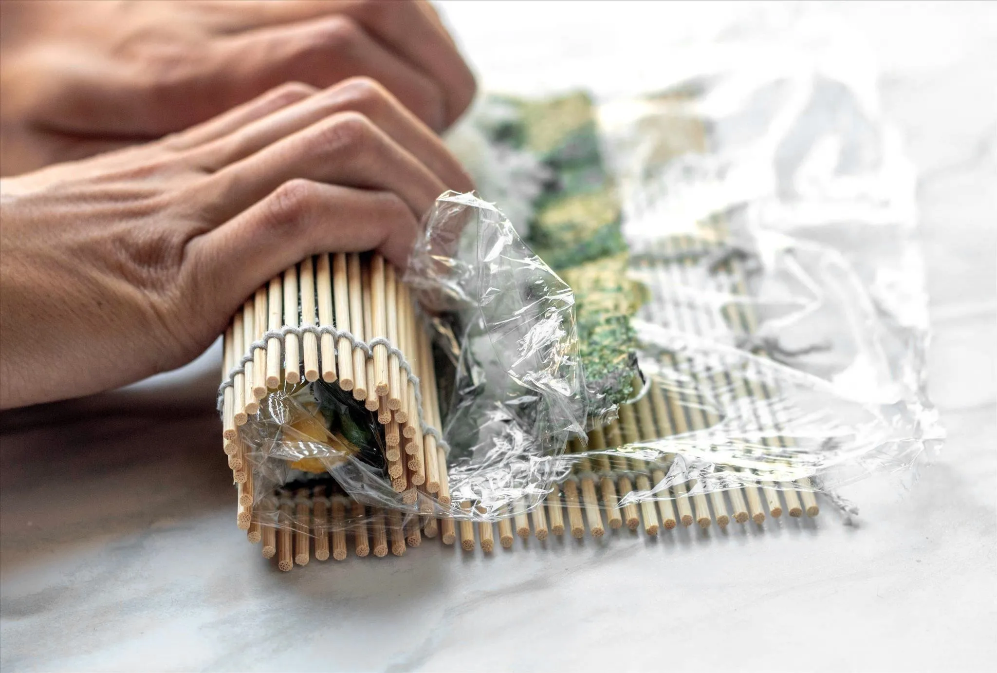 Hand preparing sushi rolls in seaweed on a plastic-wrap-lined bamboo mat.