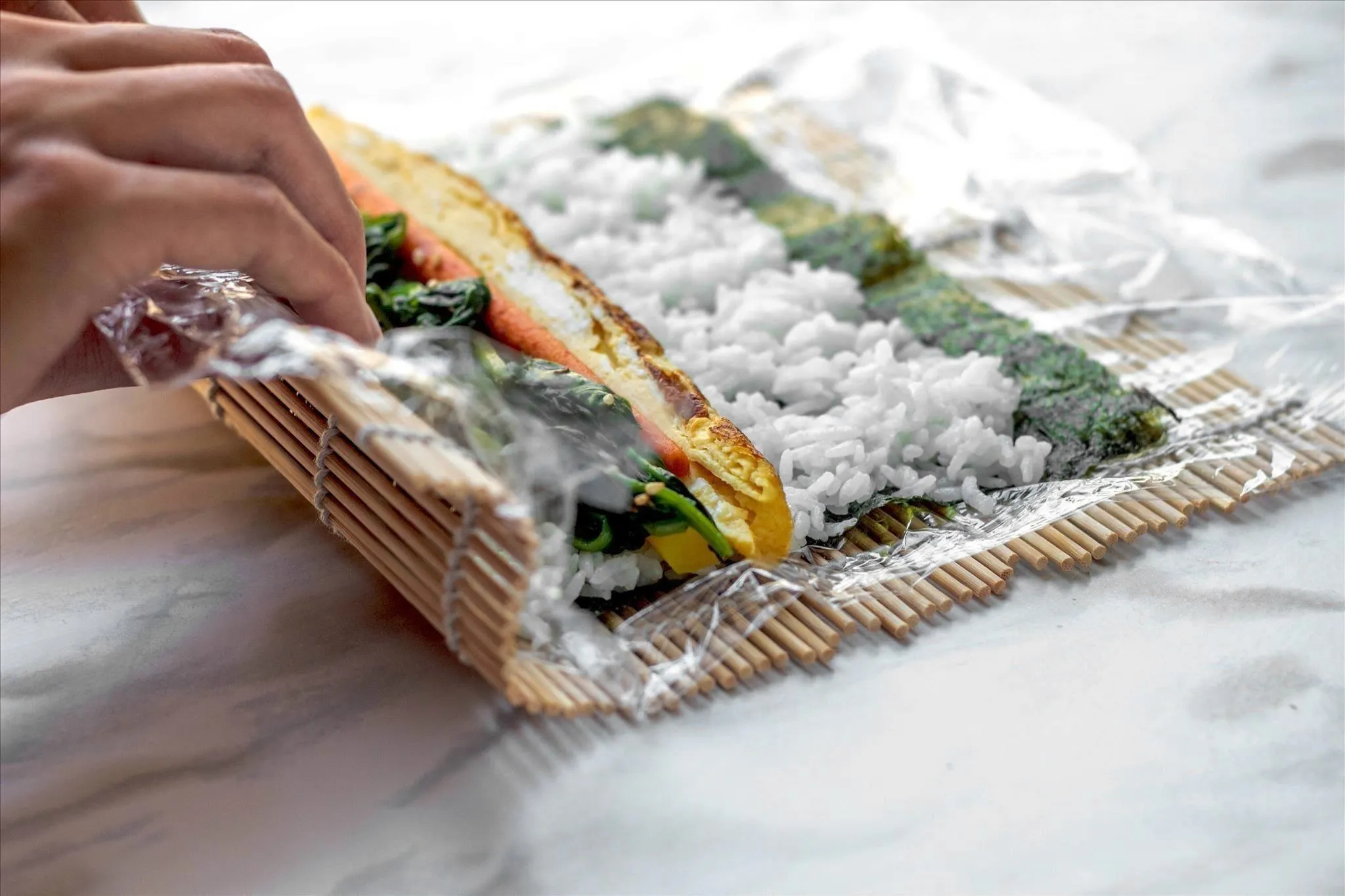 Hand preparing sushi rolls with rice and vegetables on a plastic-wrap-lined bamboo mat.
