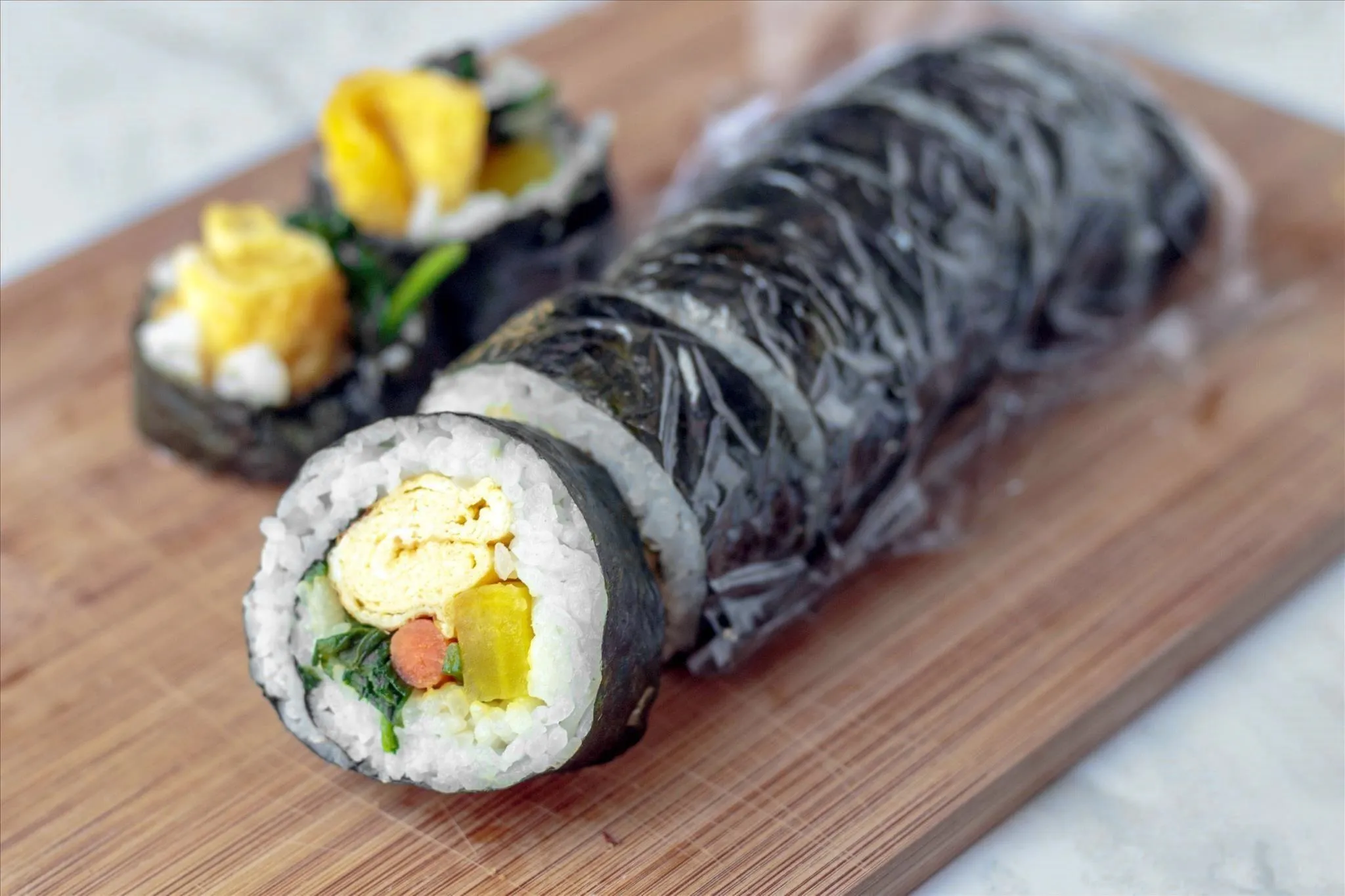 Sushi rolls with rice, tamago, spinach, burdock root, pickled daikon radish, and seaweed shell, some wrapped in plastic wrap, on a wooden cutting board.