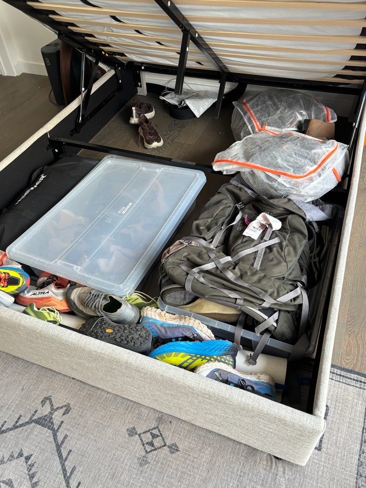Assorted shoes, bags, and clothing in the storage space underneath a lift-up compartment in a bed frame.