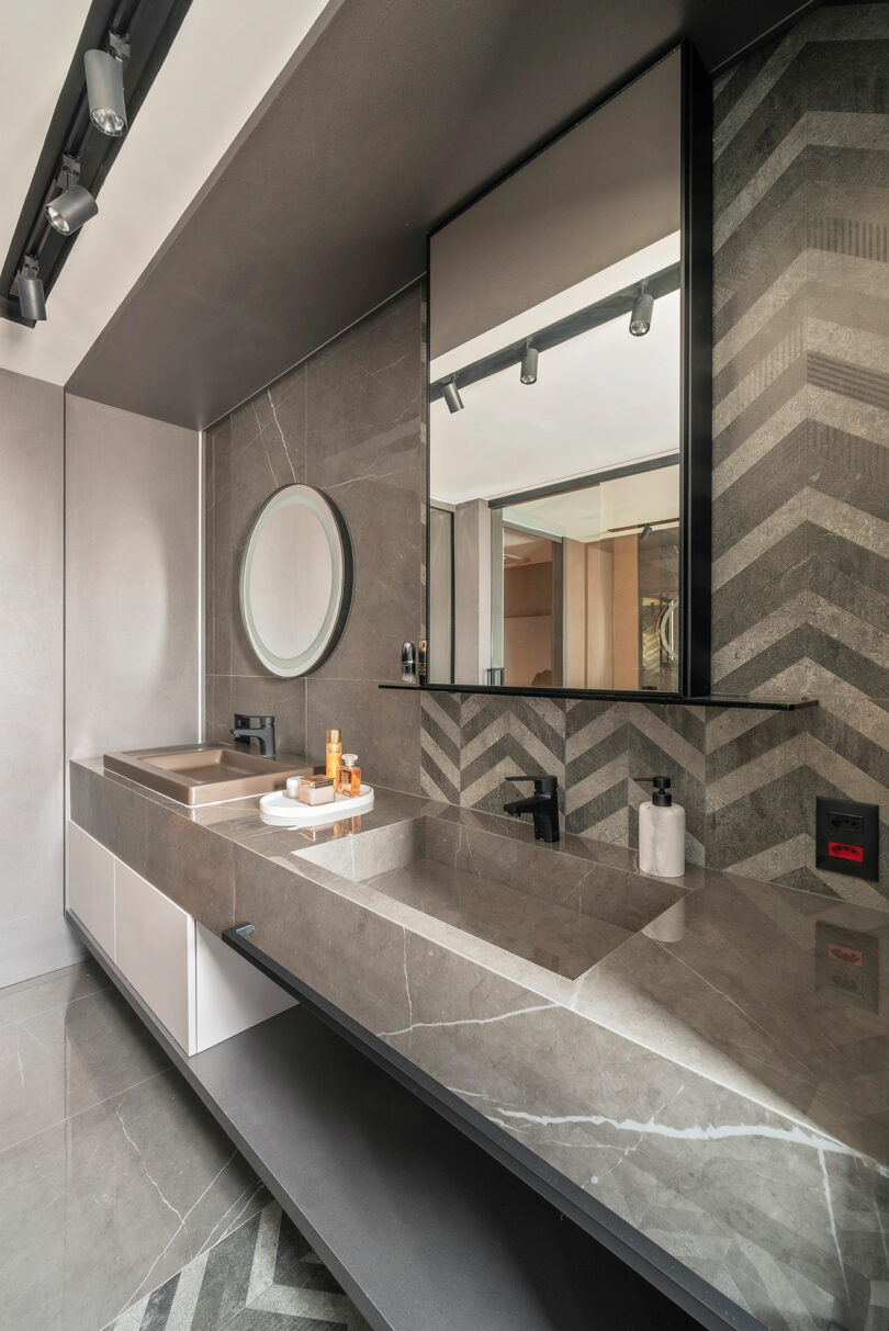 Modern bathroom with a large marble vanity, two sinks, a round mirror, and a rectangular mirror, set against gray chevron-patterned tiles and track lighting above.