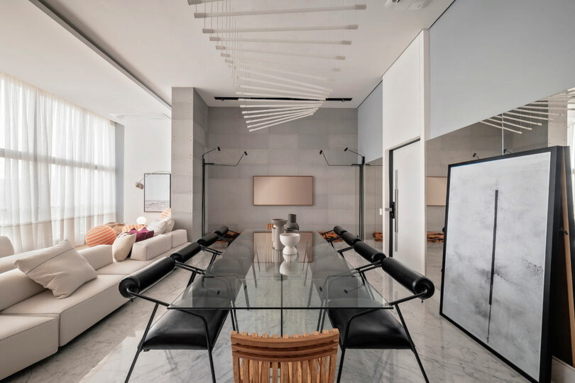 A modern dining room with a glass table, black chairs, a white sofa, abstract art, and large windows with sheer curtains letting in natural light.