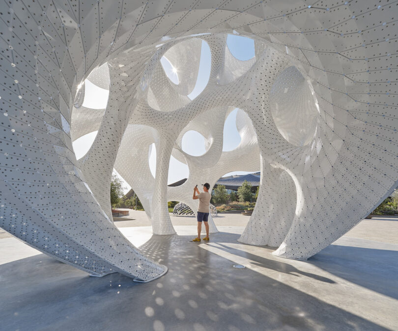 A person stands inside a large, perforated white sculptural structure with circular openings, taking a photo on a sunny day