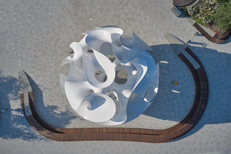 Aerial view of a large, white, abstract sculpture with swirling shapes on a paved plaza, surrounded by curved wooden benches