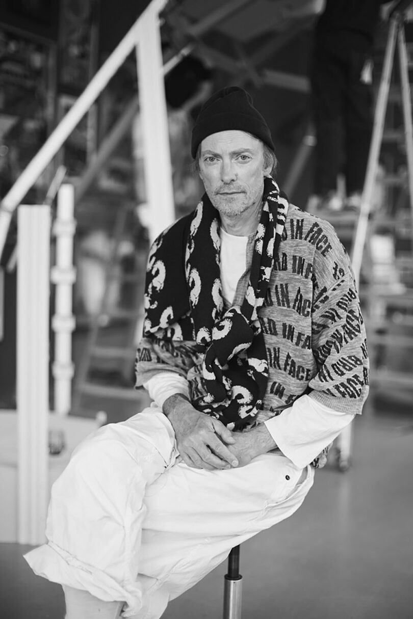 A man in a beanie and patterned scarf sits on a stool indoors, dressed in light-colored, casual clothing.