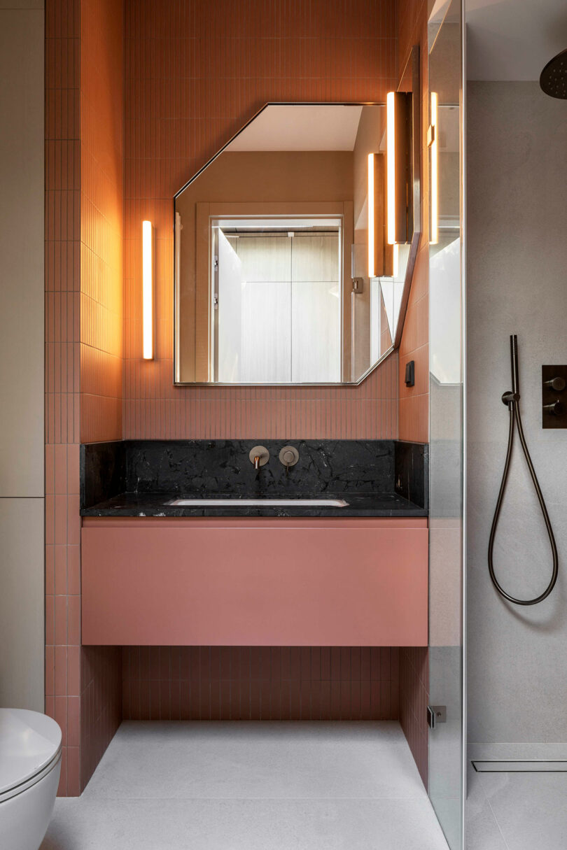 Modern bathroom with a pink vanity, black countertop, geometric mirror, wall lights, and a glass shower with a handheld showerhead.
