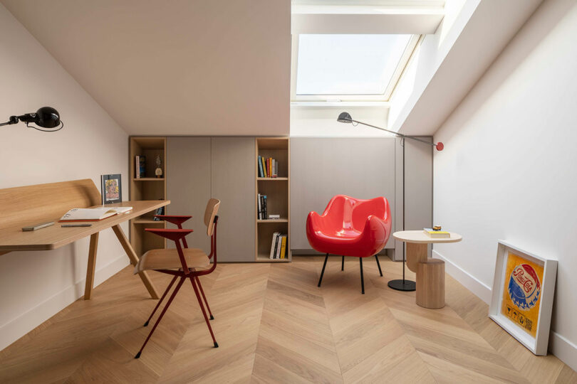 A modern attic office with a skylight, wooden desk, shelves, red armchair, and small side tables on a herringbone wood floor.