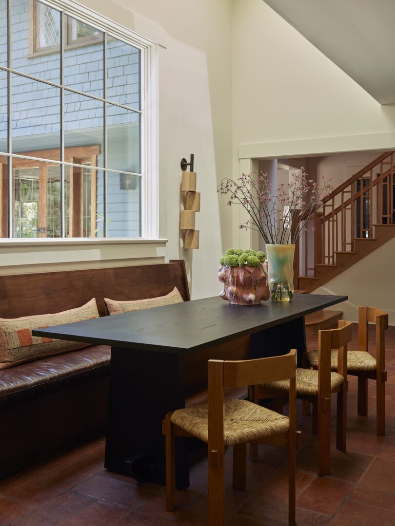 A dining area features a black rectangular table, wooden chairs with woven seats, a wooden bench, a large window, and decorative vases with flowers on the table.