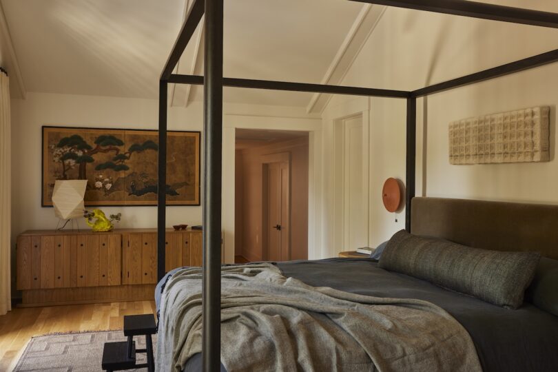 A modern bedroom with a four-poster bed, dark bedding, a wooden dresser with art and decor items, and warm natural lighting.
