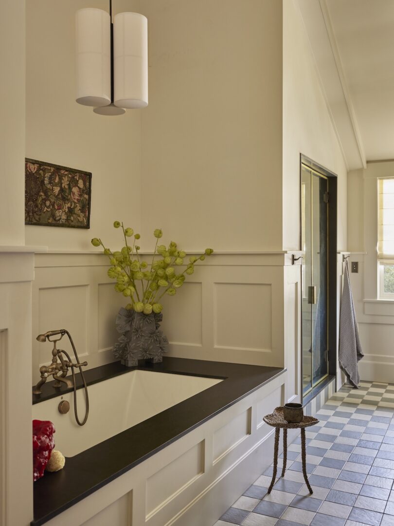 A bathroom with a built-in bathtub, black countertop, flowers in a vase, wall art, checkered tile floor, a small side table, and towels hanging by a glass shower door.