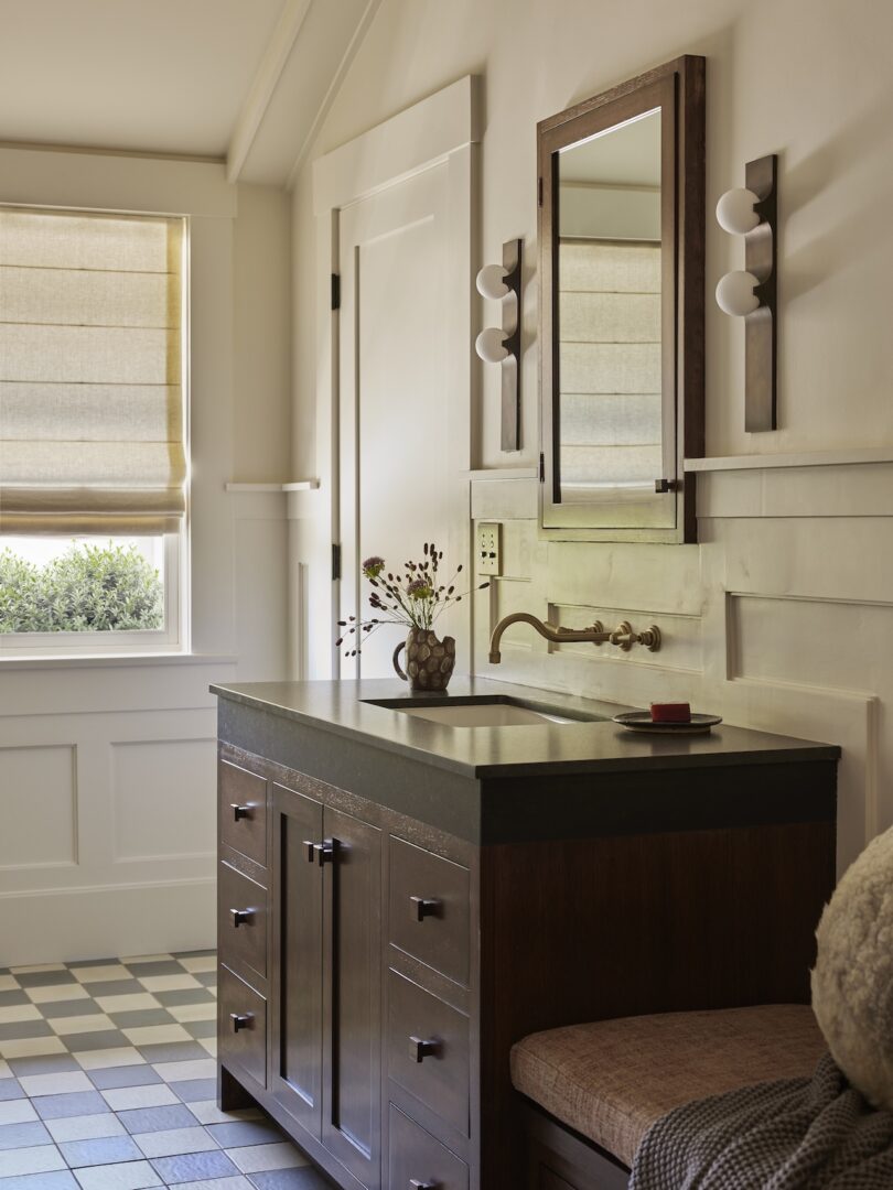 Bathroom with a dark wood vanity, wall-mounted faucet, rectangular mirror, sconce lights, checkerboard tile floor, and a window with a beige roman shade.