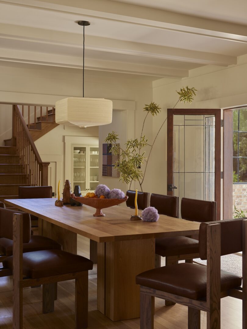 A dining room with a wooden table, six chairs, modern pendant light, and purple decorative objects. An open door and stairs are visible in the background.