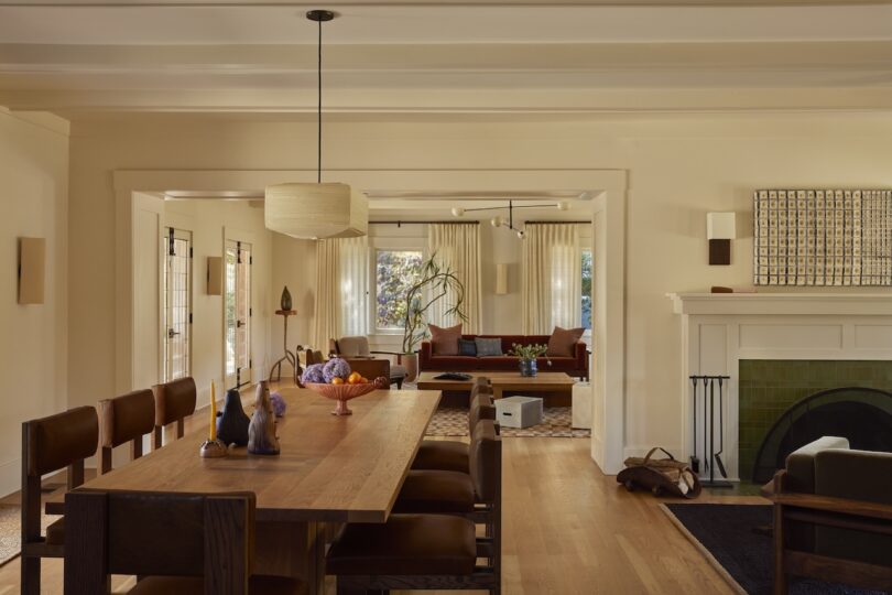 A dining room with a wooden table and chairs is adjacent to a living room with sofas, large windows, and a fireplace, all in neutral tones with natural light.