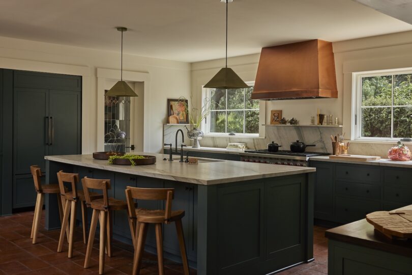 Modern kitchen with dark green cabinets, a large island with four wooden chairs, pendant lights, copper range hood, and windows providing natural light.