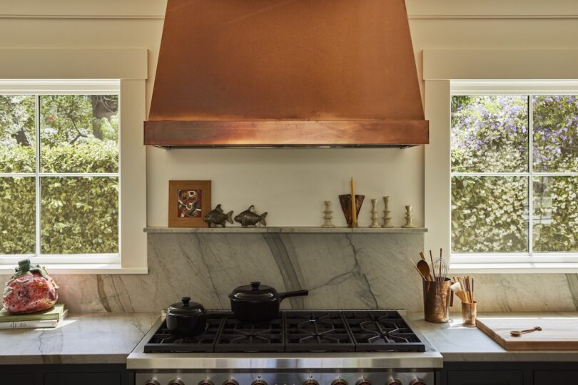 A kitchen stove with a marble backsplash, copper range hood, and windows on each side; cookware, utensils, and small decorative items are on the counter.
