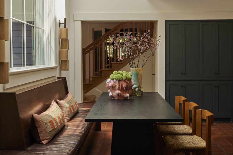 A dining area with a dark wood table, bench seating with cushions, wooden chairs, vases with flowers, and a view of stairs and cabinets in the background.