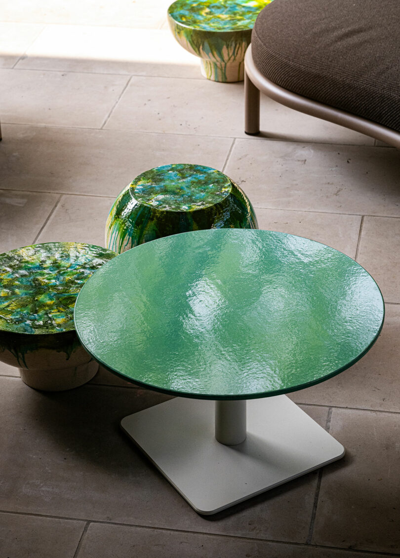A round green glass table with a white base is placed on a stone tile floor, next to two small green and white stools and part of a cushioned seat.