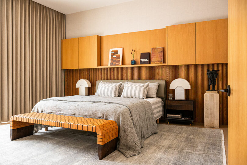 A modern bedroom with a neatly made bed, wooden wall cabinets, a woven bench, two bedside tables with lamps, and neutral-toned decor accents.