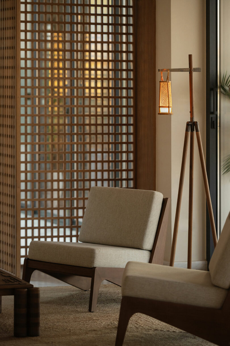 Two modern wooden chairs with beige cushions are placed near a floor lamp and in front of a wooden lattice divider in a softly lit room.