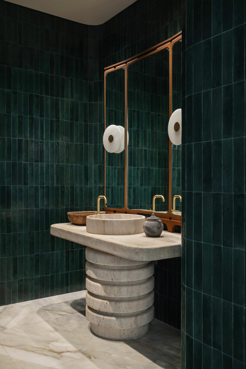 Modern bathroom with dark green tiled walls, a unique round stone sink on a sculptural vanity, two mirrors, and brass faucets.