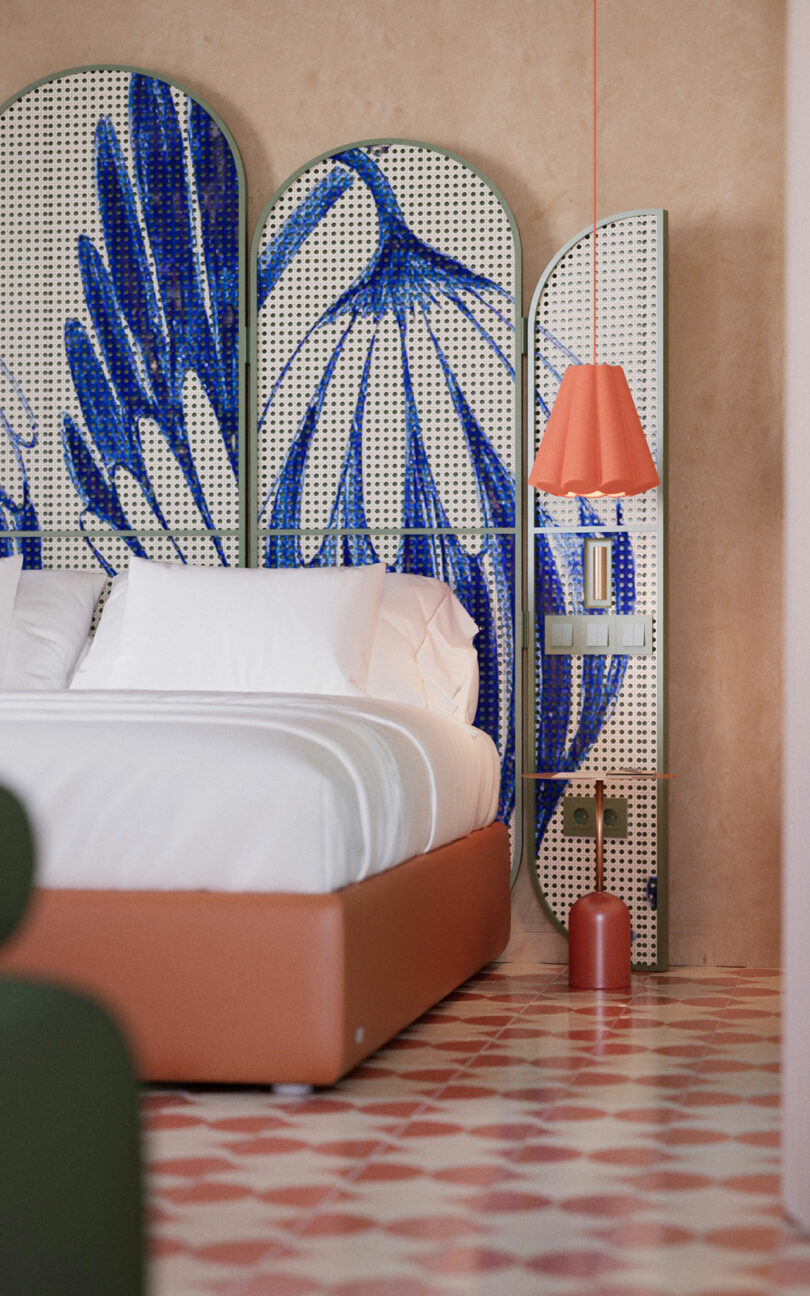 A modern bedroom with a white bed, a patterned room divider featuring blue abstract art, a red bedside lamp, and geometric tiled flooring.