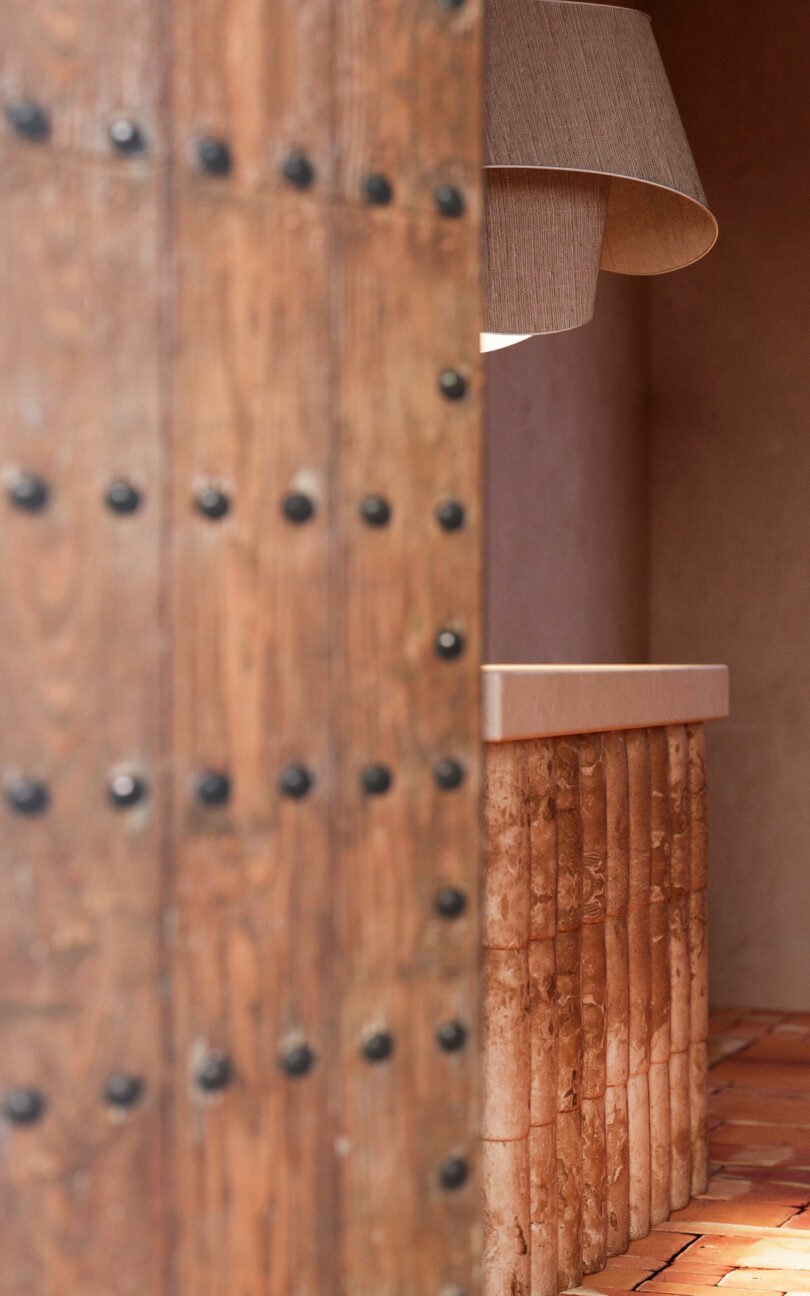 Wooden door with black rivets partially obscures a view of a textured counter, terracotta tiles, and a beige lamp in a warmly lit interior space.