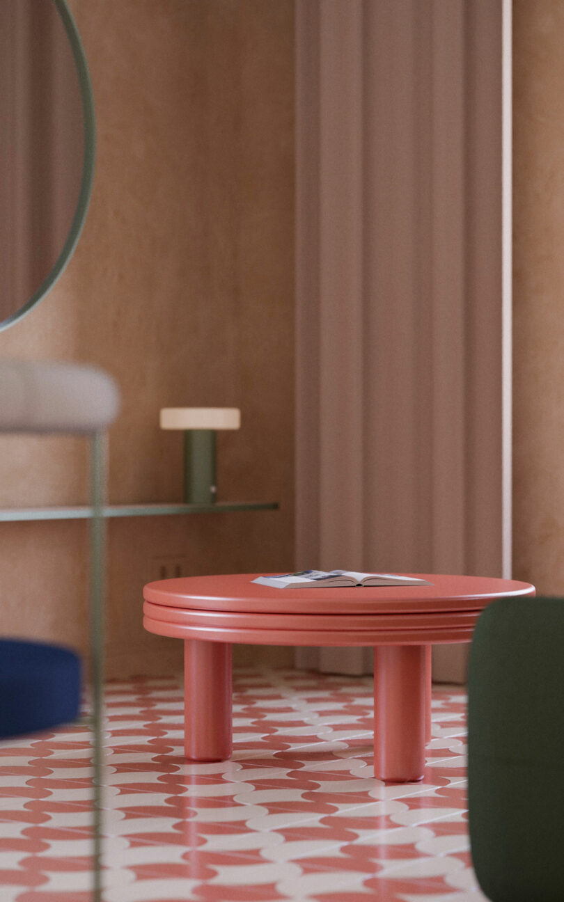 A modern room with a round pink table, patterned pink and white floor, round mirror, glass shelf, and beige curtains. An open magazine sits on the table.