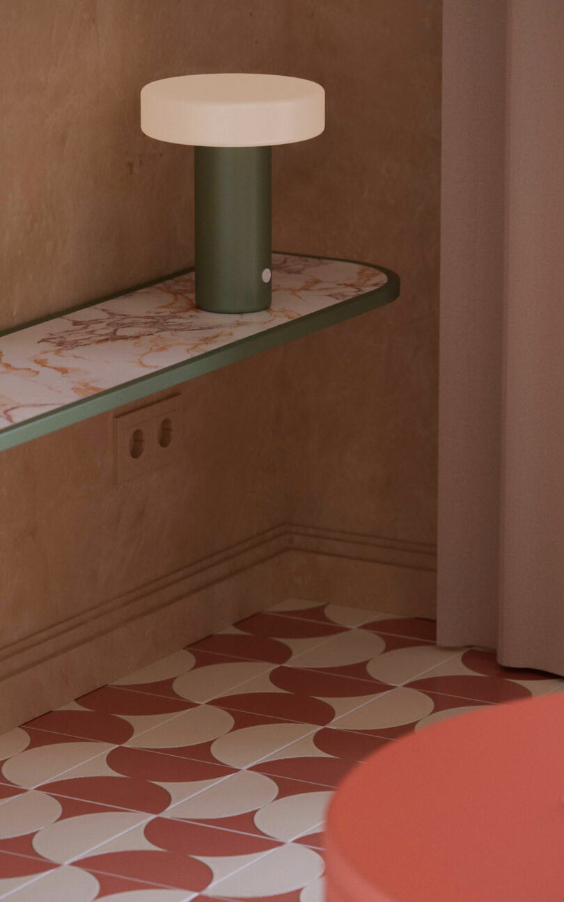 A round, modern table lamp with a green base and white shade sits on a marble shelf against a beige wall, above patterned red and white floor tiles.