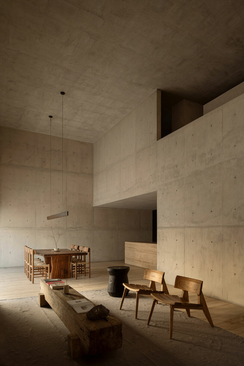 A minimalist interior with concrete walls, wooden furniture, a dining table with chairs, lounge chairs, a bench, and soft natural light.