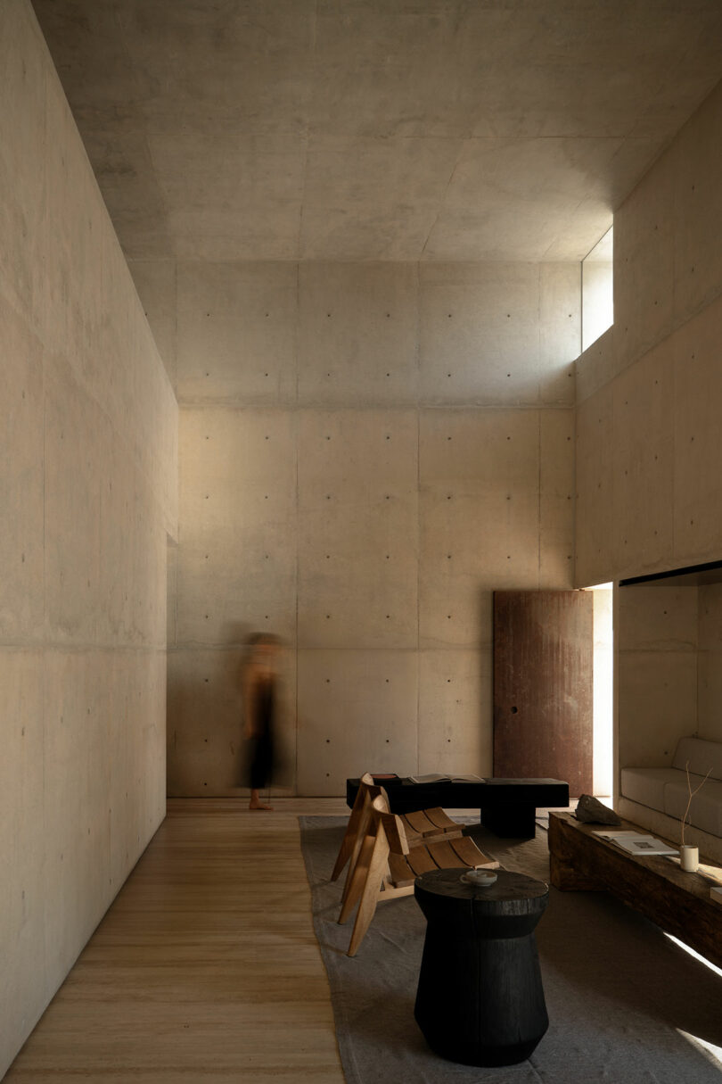 Minimalist room with concrete walls, wooden furniture, and neutral tones; a blurred person stands near the corner, and natural light enters through a high window.